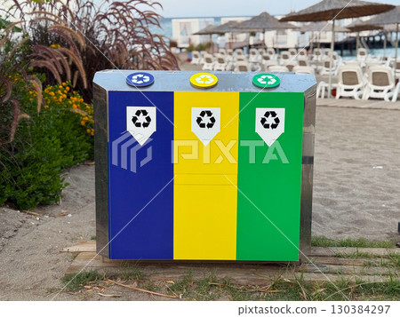 Recycling bins in blue, yellow and green on a sandy beach near umbrellas and chairs. Sustainability, environmental awareness and eco friendly lifestyle. 130384297