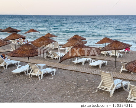 Beach with straw umbrellas and sun loungers by the sea. Summer vacation, relaxation and coastal leisure lifestyle. 130384311