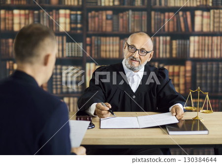 Judge explaining legal document to client at desk in library, pointing with pen 130384644