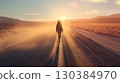 Young woman walking alone on a long straight desert road at sunset with the sun low on the horizon, beautiful golden light and dust and sand blowing across the road 130384970