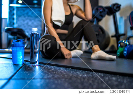 A woman taking a break and wiping sweat with a towel. A cool woman exercising at the gym. Hydration. Protein. 130384974