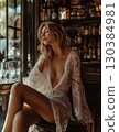 Sensual blonde woman in elegant white lace lingerie is sitting on a stool, relaxing with closed eyes in a vintage bar with a glass of wine nearby, enjoying a moment of tranquility 130384981