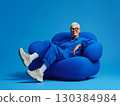 Fashionable senior woman with short gray hair and glasses smiles while relaxing comfortably on a modern, plush blue armchair against a vibrant blue backdrop, exuding confidence and style 130384984