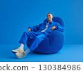 Fashionable man dressed in blue clothing and beige sneakers, sitting comfortably on a blue armchair in a vibrant blue monochrome room, smiling brightly while looking at the camera 130384986