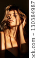 Fashion model posing with closed eyes and hand on hair, illuminated by sunlight filtering through window blinds, creating a striped shadow pattern on her face and body 130384987