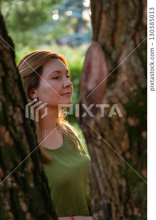 A woman in a green linen top looks upward with a serene expression, framed by tree trunks in soft evening light A woman in a green linen top looks upward with a serene expression, framed by tree trunks in soft evening light 130385013