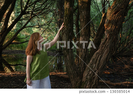 A woman in a green blouse touches a tree trunk and looks upward in a forest 130385014