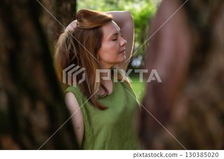 A woman in a green linen top stands among trees, her hand lifting her hair in the warm evening light A woman in a green linen top stands among trees, her hand lifting her hair in the warm evening light 130385020