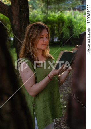 A woman in a green linen blouse reads a vintage book outdoors, framed by trees in soft evening light A woman in a green linen blouse reads a vintage book outdoors, framed by trees in soft evening light 130385035