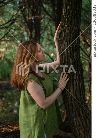 A woman in a green blouse touches a tree trunk and looks upward in a forest 130385038