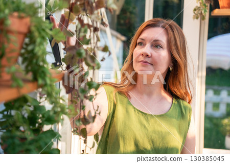 Woman in a green linen top gently touches hanging plants inside a bright greenhouse, with natural light highlighting her face 130385045