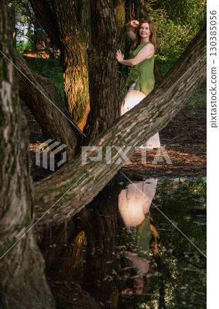 A woman in a green blouse and white trousers stands among tree trunks, her reflection visible in the water below A woman in a green blouse and white trousers stands among tree trunks, her reflection visible in the water below 130385056