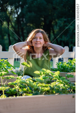 Woman in a green linen top sits among raised garden beds with strawberry plants, smiling in the sunlight 130385057