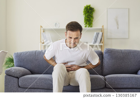 Young man hunched on couch with abdominal pain, pressing stomach in discomfort 130385167
