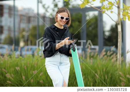 Stylish young woman with smartphone scans QR code for electric scooter rental. Girl using app on phone to unlock dockless transport on street. Urban style riding ecological vehicle. Modern technology Stylish young woman with smartphone scans QR code for electric scooter rental. Girl using app on phone to unlock dockless transport on street. Urban style riding ecological vehicle. Modern technology 130385728