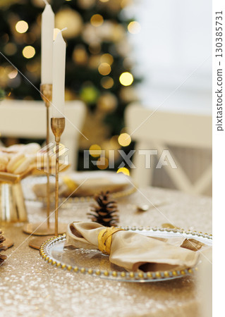 Luxury golden Christmas dinner table setting. Glowing tablecloth, napkin with steel napkin ring on glass plate, cutlery, candlesticks. Xmas tree illuminating on backdrop. Festive new 2026 year party. Luxury golden Christmas dinner table setting. Glowing tablecloth, napkin with steel napkin ring on glass plate, cutlery, candlesticks. Xmas tree illuminating on backdrop. Festive new 2026 year party. 130385731