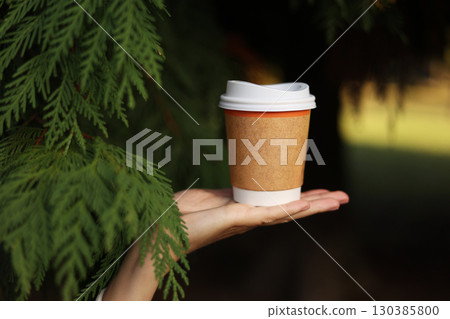 Hand holds eco zero waste paper cup in leaves of white cedar, white plastic recycle lid take away with hot drink, coffee or tea outdoors in park, place for text. Seasonal warm drinks to go in cold day 130385800