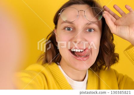 Playful young woman takes a selfie, smiles, sticks out her tongue, holds glasses on yellow backdrop. Modern lifestyle, social media, youth influencer marketing, self expression, personal branding 130385821