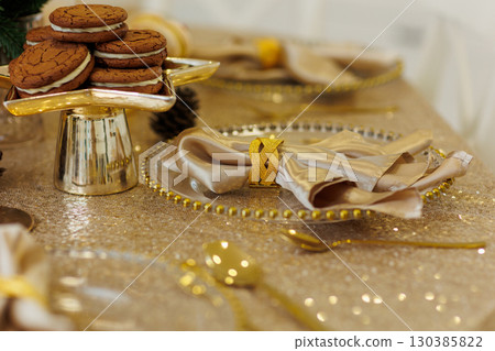 Luxury golden Christmas dinner table setting. Glowing tablecloth, napkin with steel napkin ring on glass plate, cutlery, candlesticks. Xmas tree illuminating on backdrop. Festive new 2026 year party. 130385822