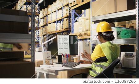 Storehouse black worker reviewing digital shipment status, using awb numbers on tracking software. Woman handling data driven order tracking and delivery updates in warehouse. Camera A. 130387260