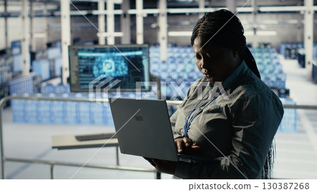 Data center admin checks AI dashboard using laptop during rigs systems review. African american woman in server hub tests artificial intelligence tools on notebook to check reliable performance 130387268