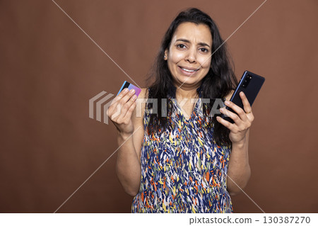 Anxious woman stares at camera, with smartphone in hand, distressed after discovering bank card has been hacked, reacting to suspected online fraud. Worried female individual holding card and phone. 130387270