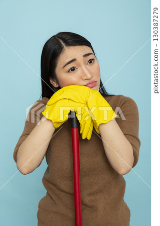 Portrait of asian female cleaner leaning on red broomstick with sad expression, feeling exhausted after long workday. Thoughtful housekeeper pauses to rest, lost in deep reflection and fatigue. 130387279
