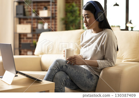 Asian woman wearing wireless headphones and holding tea mug, attentively watching videos on laptop. Female entrepreneur seated in cozy brick wall apartment, taking break from remote work tasks. 130387409