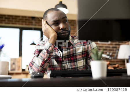 African american male entrepreneur shows low energy, lack of motivation and disinterest in business tasks. Tired remote worker sitting at desk, holding head with hand and staring at desktop monitor. African american male entrepreneur shows low energy, lack of motivation and disinterest in business tasks. Tired remote worker sitting at desk, holding head with hand and staring at desktop monitor. 130387426