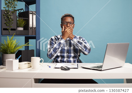 Black male entrepreneur seated at office desk, covering his mouth, showing a sign of no sharing of information. African american businessman with glasses, places his hands on face and looks at camera. 130387427