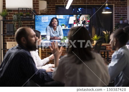 Multiethnic colleagues discussing startup project plans in late night meeting. A video conference with remote team is visible on digital screen, symbolizing modern business collaboration. Multiethnic colleagues discussing startup project plans in late night meeting. A video conference with remote team is visible on digital screen, symbolizing modern business collaboration. 130387428