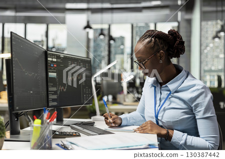 Risk analyst in office documents strategies while observing stock candle patterns on screen. Brokerage company worker reviews stock market shares volatility, recording insights in notepad 130387442