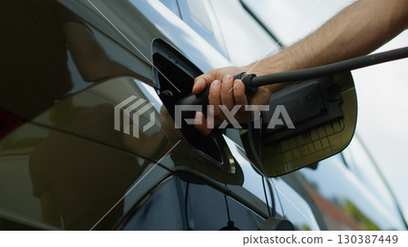 Close up of driver recharging EV vehicle battery powering sustainable electric motor, low angle shot. Person recharges battery of EV car by plugging into clean energy power source before long trip Close up of driver recharging EV vehicle battery powering sustainable electric motor, low angle shot. Person recharges battery of EV car by plugging into clean energy power source before long trip 130387449