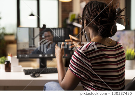 Black woman uses desktop pc for virtual meeting, discussing marketing strategies with business advisor from home. Male consultant appears on screen, offering strategic insights to focused entrepreneur Black woman uses desktop pc for virtual meeting, discussing marketing strategies with business advisor from home. Male consultant appears on screen, offering strategic insights to focused entrepreneur 130387450