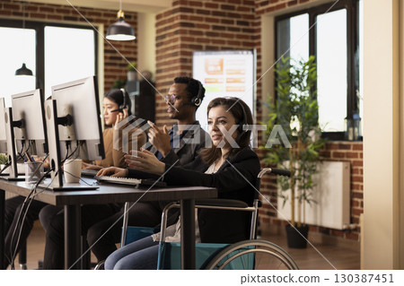 Smiling female employee, wearing wired headphones, works diligently at her desk. Caucasian woman in wheelchair uses her headset to communicate with online clients, entering data into her computer. Smiling female employee, wearing wired headphones, works diligently at her desk. Caucasian woman in wheelchair uses her headset to communicate with online clients, entering data into her computer. 130387451