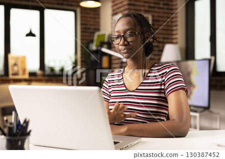 African american entrepreneur with glasses seated at desk, engaging in virtual training session with colleagues on laptop. Remote businesswoman participates in professional webinar from home office. African american entrepreneur with glasses seated at desk, engaging in virtual training session with colleagues on laptop. Remote businesswoman participates in professional webinar from home office. 130387452