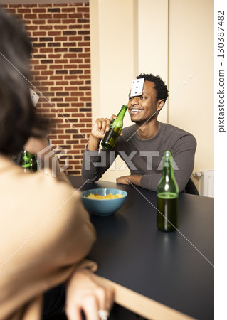 Smiling african american man sitting at table and holding beer bottle, casually hanging out with friends at home. Black male individual with card on forehead, playing guessing game in brick wall room. 130387482
