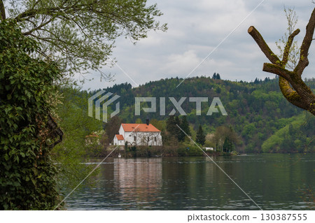 White church with red roof by forested riverbank - peaceful spring landscape with cloudy sky 130387555