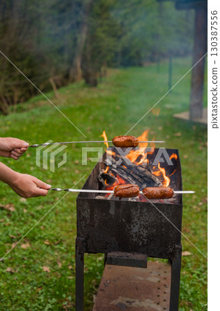 Grilling sausages on skewers over open flame in metal barbecue grill during outdoor cookout . 130387556