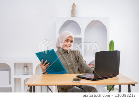 Smiling Young Woman Working on Laptop with Folders in Cozy Office Space 130388212