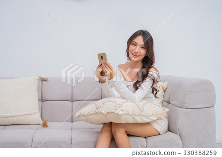 A Cheerful Young Woman Engaged in Using Her Smartphone While Relaxing on a Cozy Couch 130388259