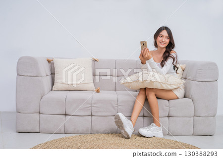 A Young Woman Resting Comfortably on a Couch while Holding Her Phone with a Bright Smile 130388293