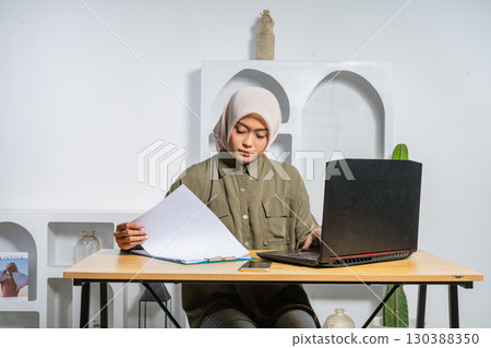 Young Woman Analyzing Documents at a Desk with Laptop in Modern Workspace Young Woman Analyzing Documents at a Desk with Laptop in Modern Workspace 130388350