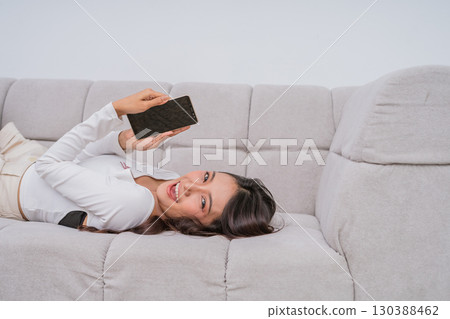 A young woman is happily relaxing on a comfortable couch while using her smartphone A young woman is happily relaxing on a comfortable couch while using her smartphone 130388462