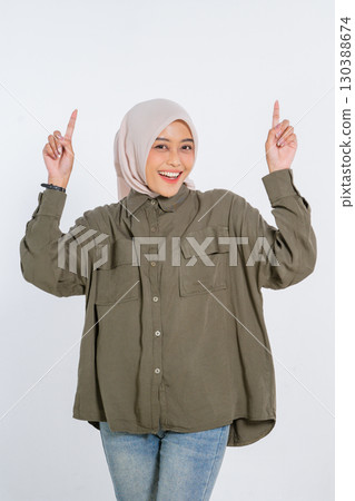 A Cheerful Young Woman in an Olive Shirt and Hijab is Pointing Upward Excitedly A Cheerful Young Woman in an Olive Shirt and Hijab is Pointing Upward Excitedly 130388674