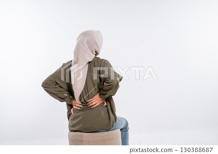 A woman wearing a hijab is sitting in a chair while experiencing back pain and discomfort 130388687