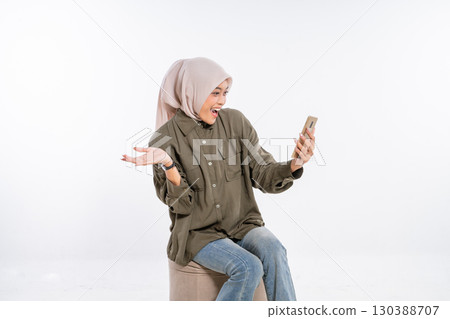 A joyful and excited woman wearing a hijab is engaging with her smartphone device 130388707