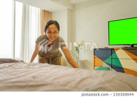 Cheerful young woman happily making her bed in a modern bedroom featuring a TV Cheerful young woman happily making her bed in a modern bedroom featuring a TV 130389044