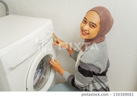 A woman wearing a hijab is skillfully using a washing machine within her home 130389081