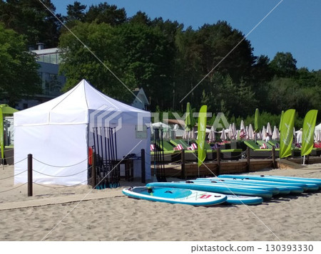 Sandy beach area with a row of blue stand-up paddleboards lined up near a white canopy tent. Paddles are propped against a wooden post. Yurmala beach, Latvia  130393330